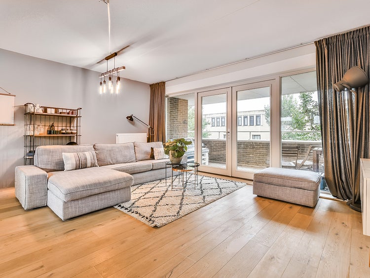 Helles Wohnzimmer mit grauem Ecksofa, passendem Hocker, hellem Holzfußboden, gemustertem Teppich und großen Glastüren zum Balkon.