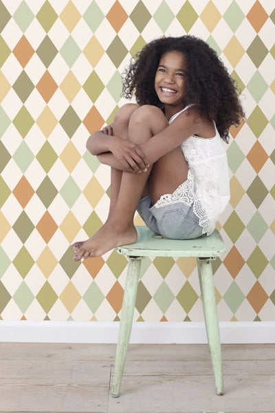 Mädchen sitzt auf einem Hocker vor einer Tapete mit Rautenmuster.