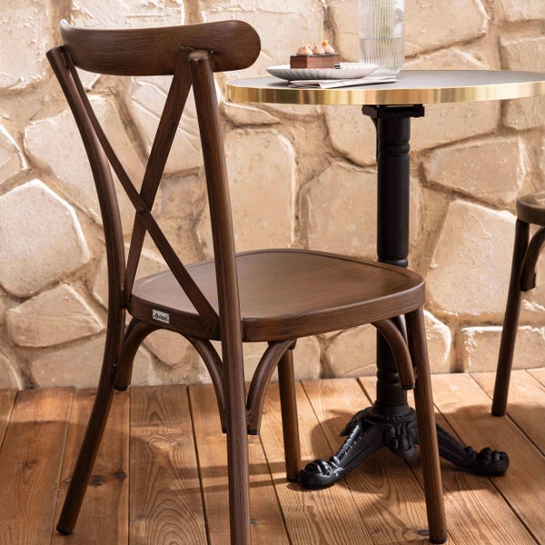 Bistrostuhl in dunkler Holzoptik mit Kreuzruecken an einem runden Tisch auf einer Terrasse mit Holzboden und Steinwand.