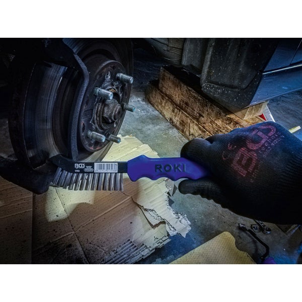 Eine Hand mit Handschuh reinigt mit einer Drahtbürste eine Bremsscheibe am Auto.