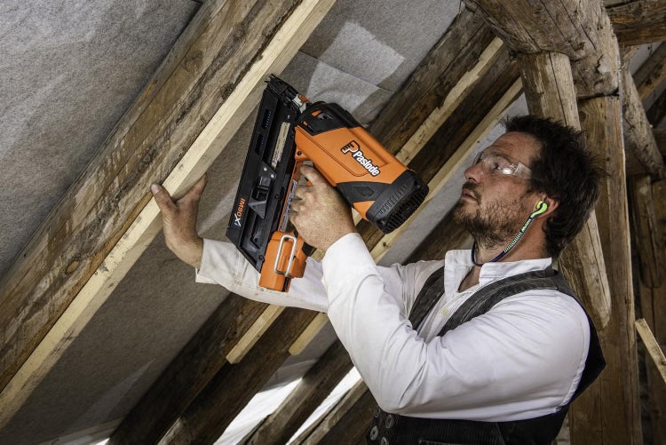 Ein Handwerker nutzt einen Paslode gasbetriebenen Streifennagler bei Dacharbeiten an einem Holzdachstuhl mit Schutzbrille und Gehörschutz.