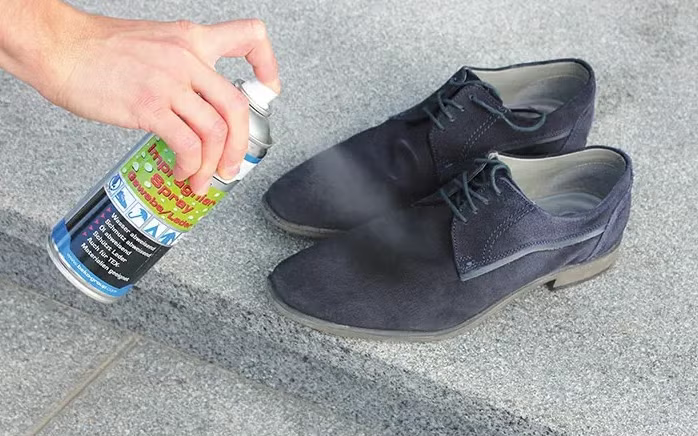 Hand beim Auftragen von Imprägnierspray auf ein Paar dunkle Wildlederschuhe zum Schutz vor Wasser und Schmutz.