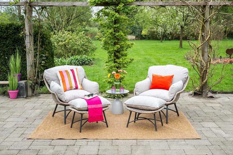 Zwei bequeme Garten-Loungesessel mit Hockern, Polstern und bunten Kissen auf einem Außenteppich unter einer Pergola im Garten.