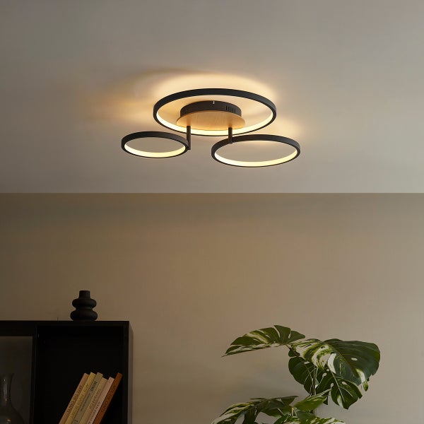 Moderne LED Deckenleuchte mit drei ringförmigen Elementen aus Metall und Holzelementen in einem Wohnraum.
