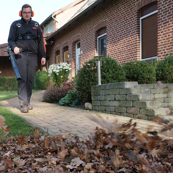 Mann mit Laubbläser räumt Laub vor einem Backsteinhaus