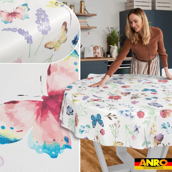 Runde Tischdecke mit bunten Schmetterlingen und Blumenmustern in einer hellen Küche. Eine Frau streicht das Tischtuch auf einem Tisch glatt. ANRO Logo.