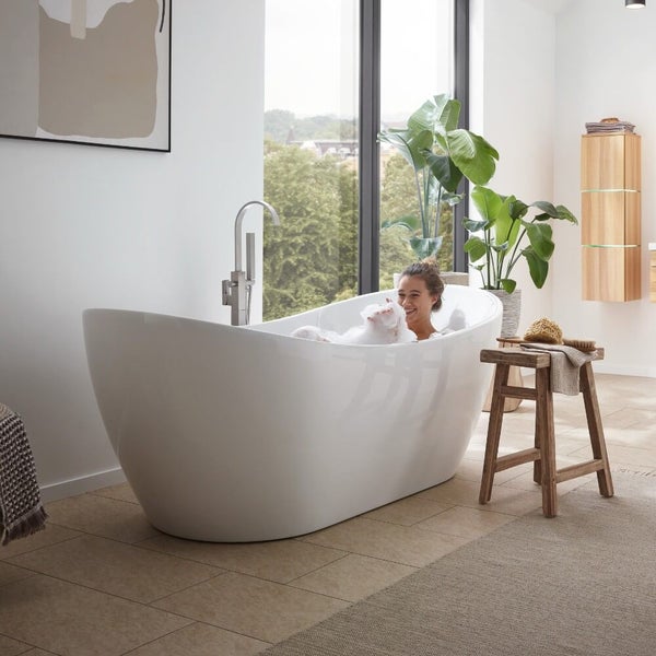 Freistehende Badewanne in einem hellen Badezimmer mit einer Frau im Schaumbad, daneben ein Holzhocker und eine große Zimmerpflanze am Fenster.
