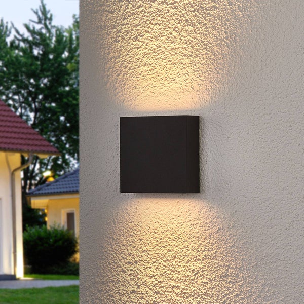 Schwarze quadratische Außenwandleuchte mit Licht nach oben und unten an einer verputzten Fassade, im Hintergrund ein Wohnhaus mit Garten.