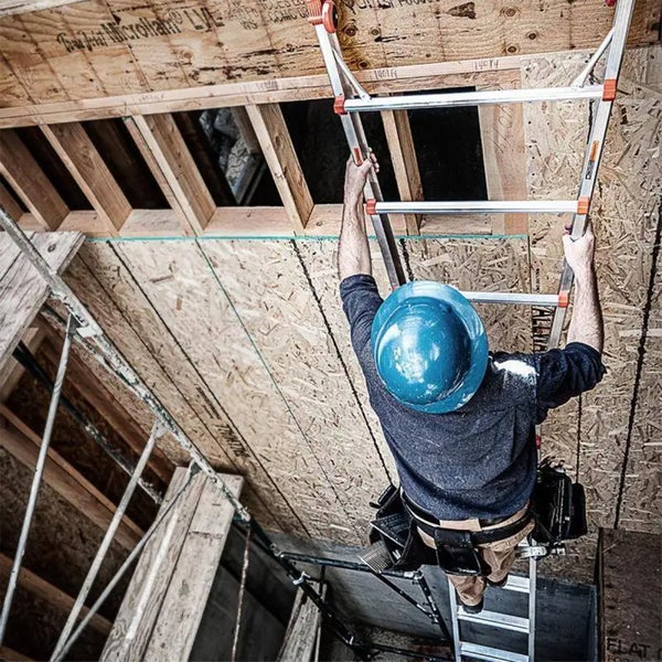 Handwerker mit Schutzhelm steigt auf einer Aluminium Mehrzweckleiter in einem Rohbau mit Holzständerwerk.