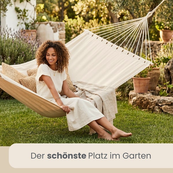 Frau entspannt in einer hellen Tuchhängematte mit Spreizstab in einem hellen, sonnigen Garten mit Rasen, Lavendel und Terracotta-Töpfen.