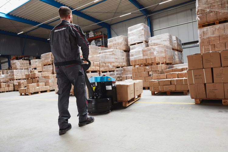 Ein Mitarbeiter bedient einen elektrischen Hubwagen in einem Lagerhaus mit vielen gestapelten Kartons auf Paletten.