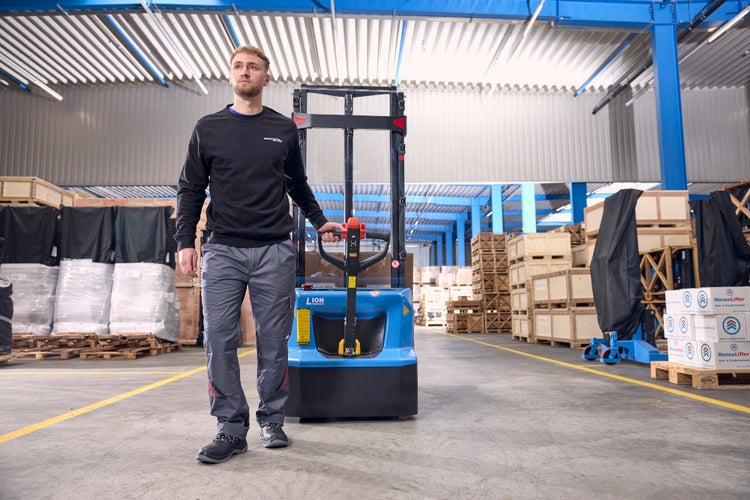 Lagerarbeiter bedient einen blauen Elektro-Hochhubwagen in einem hellen, organisierten Lager mit Holzkisten und Kartons.