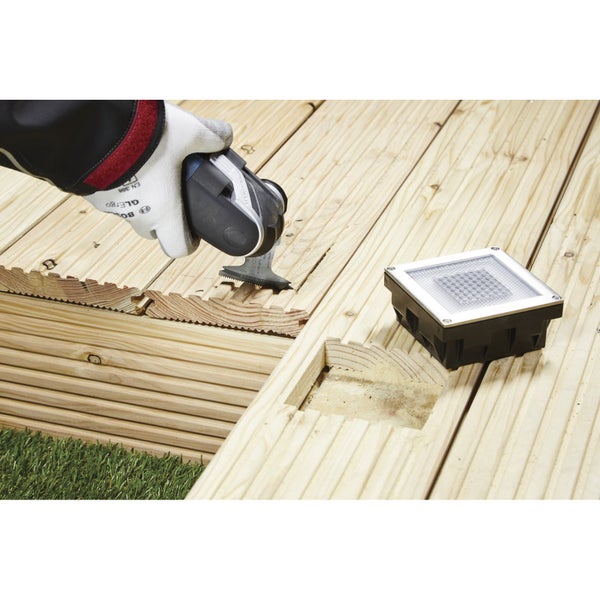 Szene mit einer Hand, die mit einem Multifunktionswerkzeug eine Aussparung in eine Holzterrasse schneidet, neben einer quadratischen Bodenleuchte.