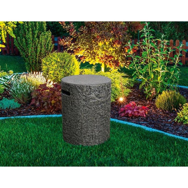 Zylindrisches Gartenelement in Natursteinoptik auf einer Rasenfläche in einem beleuchteten Garten mit Sträuchern und Stauden.