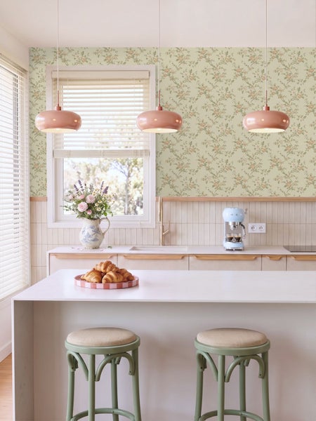 Helle Küche im Landhausstil mit floraler Tapete, Kücheninsel, zwei Barhockern, drei Pendelleuchten und Arbeitsplatte mit Spüle und Kaffeemaschine.