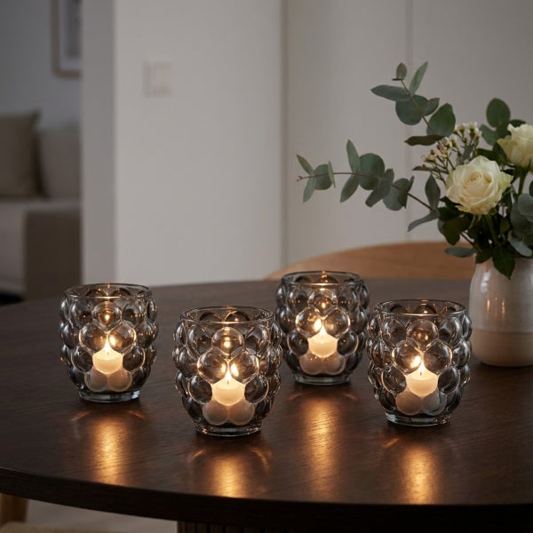 Vier Teelichthalter aus Glas mit strukturierter Oberfläche auf einem dunklen Holztisch mit einer Blumenvase im Hintergrund.