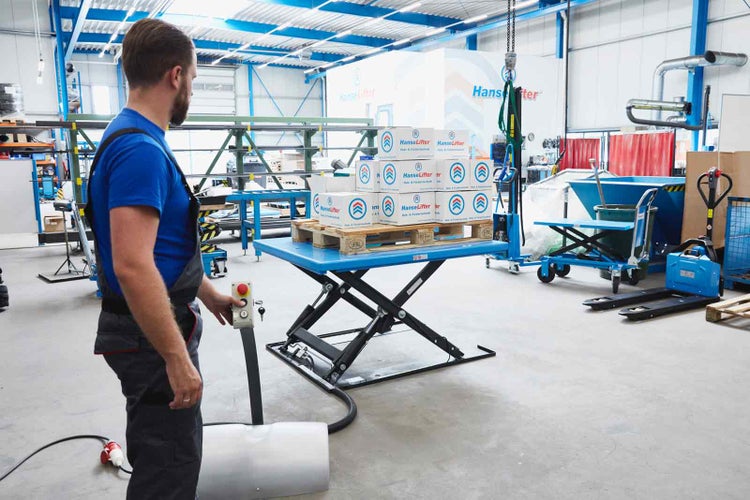 Ein Mann bedient einen blauen stationären Scherenhubtisch in einem hellen Lager, der eine Palette mit Kartons mit HanseLifter Logo trägt.