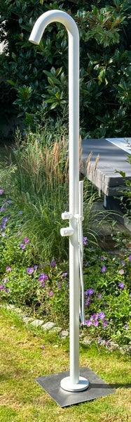 Weiße Gartenbrause mit schlanker Säule, integrierter Handbrause und quadratischer Bodenplatte in einer Gartenumgebung mit Pflanzen.