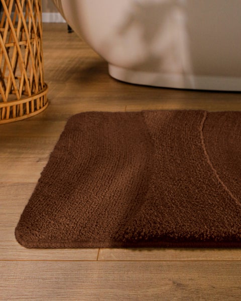 Braune Badematte mit weicher Wellenstruktur auf hellem Holzboden in einem Badezimmer neben einer freistehenden Badewanne.
