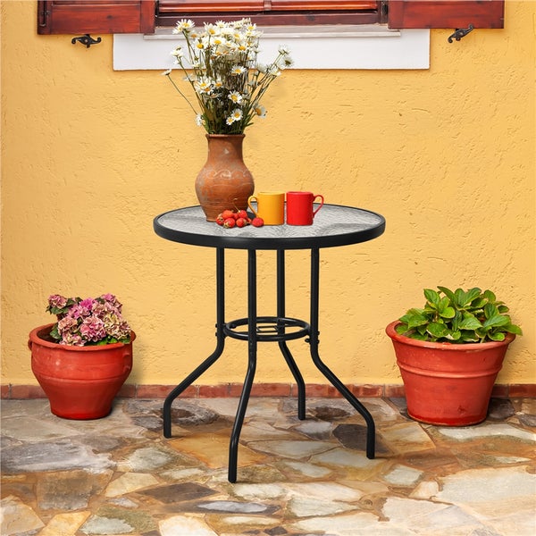 Runder Bistrotisch mit Glasplatte und Metallgestell auf einer Terrasse mit Natursteinboden, dekoriert mit Blumen und Pflanzgefäßen.