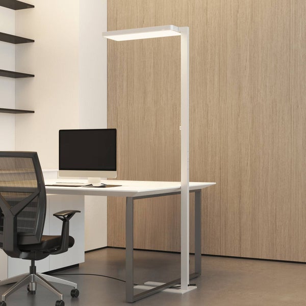 Moderne Büroumgebung mit einer hohen weißen LED-Stehleuchte neben einem Schreibtisch mit Monitor und schwarzem Bürostuhl vor einer Holzwand.