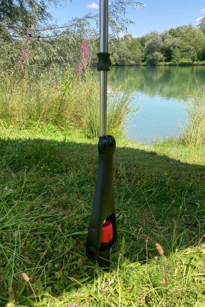 Bodenhülse aus Metall mit schwarzer Kunststoffbasis und rotem Einstellrad auf einer Wiese an einem See.