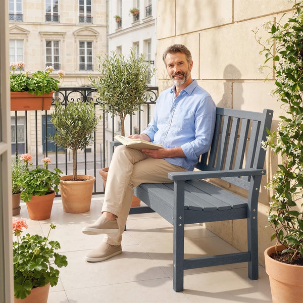 Mann sitzt auf einer Gartenbank aus Holz auf einem sonnigen Balkon, umgeben von blühenden Pflanzen und einem Ausblick auf ein städtisches Gebäude.