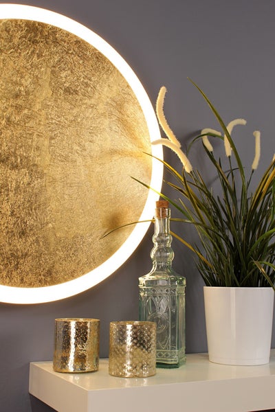 Runde LED-Wandleuchte in Goldoptik mit Leuchtring neben einer Glasflasche und einer Pflanze auf einem Regal.