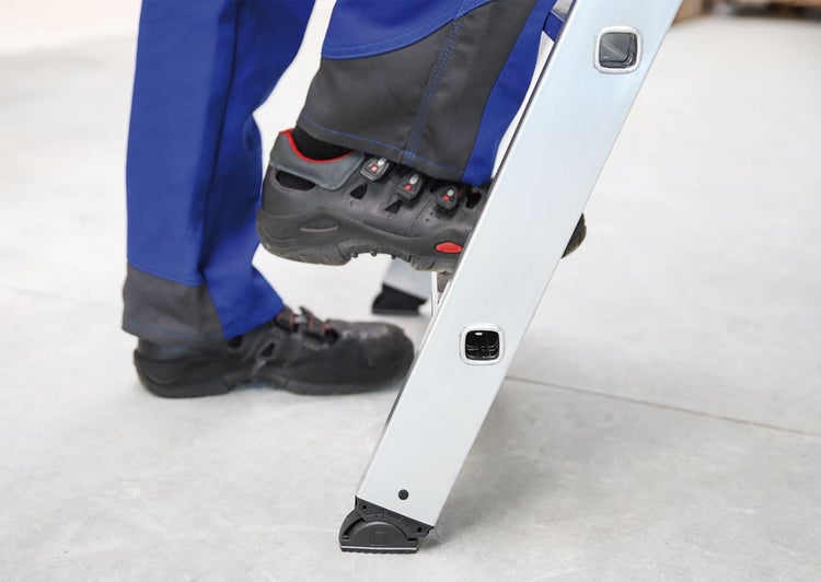 Person in Arbeitshose und Sicherheitsschuhen steht auf einer stabilen Aluminiumleiter.