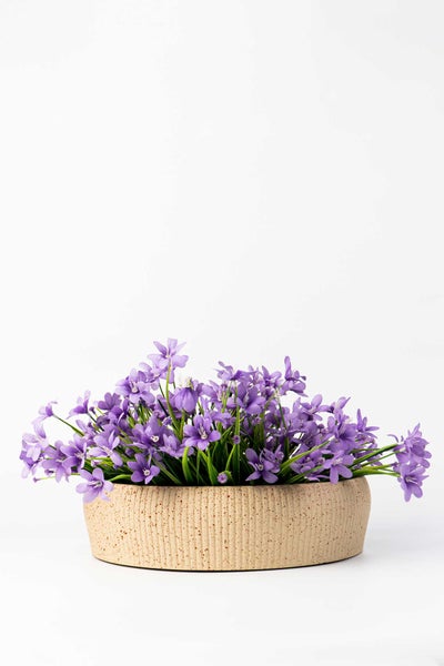 Künstliche lila Blumen in einer flachen, strukturierten beigen Dekoschale.
