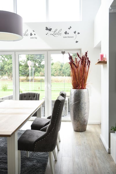 Helles Esszimmer mit Holztisch, dunkelgrauen Polsterstühlen und großer silberner Bodenvase mit hohen roten Pflanzen neben Glastüren.