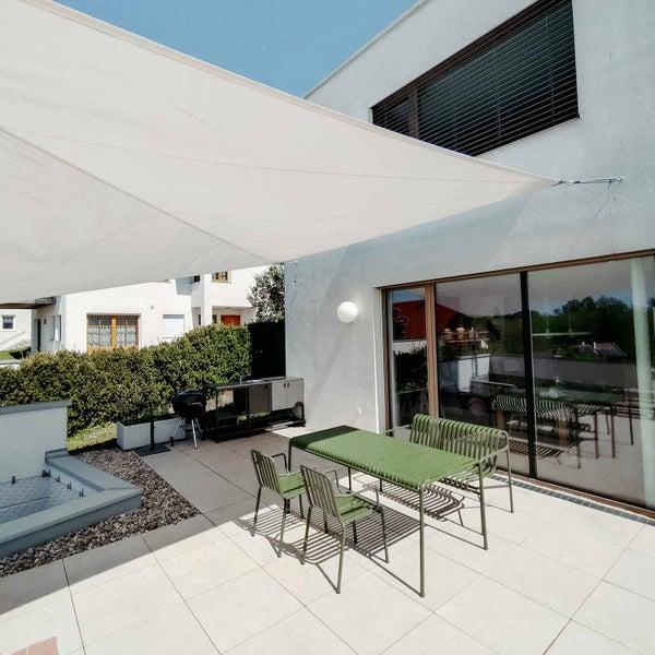Helle, modern gestaltete Terrasse mit weißem Sonnensegel, grüner Metall-Sitzgruppe und einer Außenküche vor einem Haus.