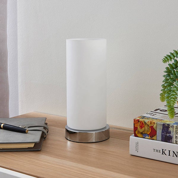 Moderne Tischleuchte mit weißem Glaszylinder und verchromtem Metallfuß auf einem Holzschreibtisch neben Notizbuch und Büchern.