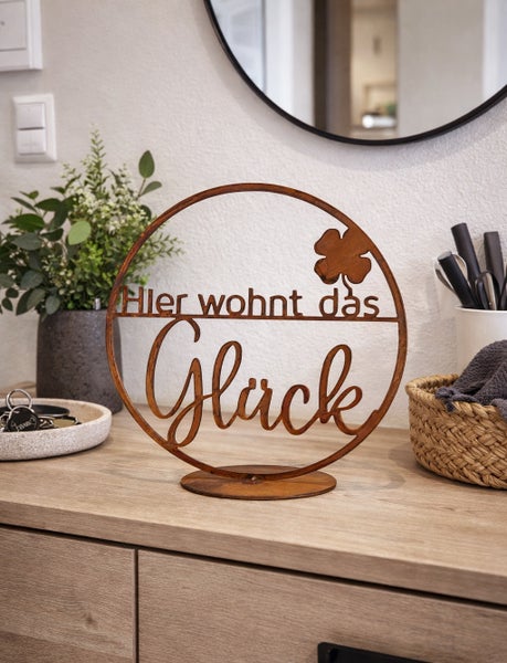 Runder Aufsteller aus Metall in Rostoptik mit dem Schriftzug Hier wohnt das Glück und einem vierblättrigen Kleeblatt auf einer Kommode.