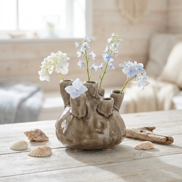 Keramikvase mit mehreren Öffnungen für Blumen, dekorativ arrangiert auf einem Holztisch mit Muscheln und Treibholz im maritim inspirierten Raum.