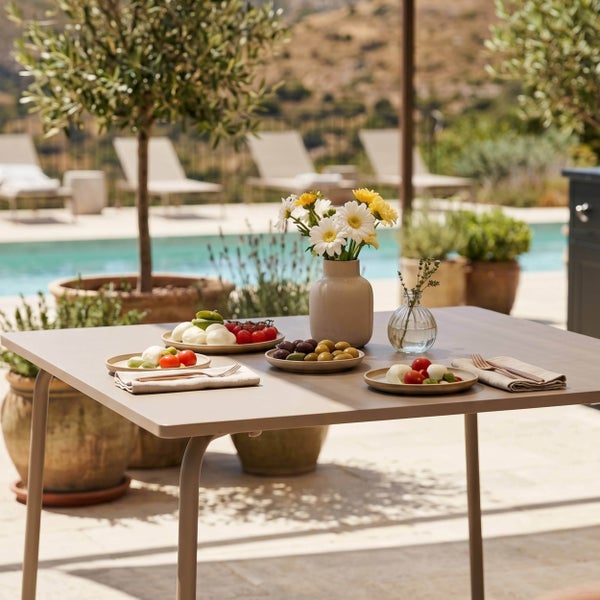 Quadratischer Gartentisch aus Metall auf einer hellen Terrasse am Pool, gedeckt mit Tellern, Blumenvasen und Besteck.