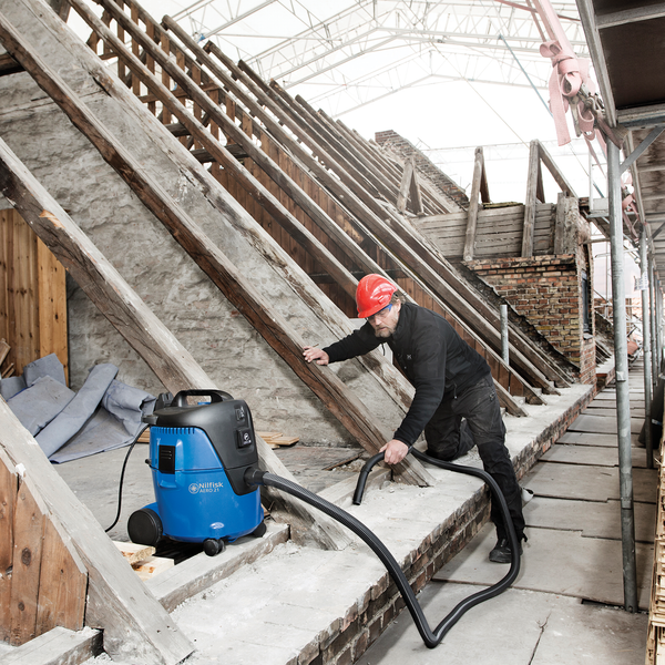 Ein Handwerker nutzt den Nilfisk Nass-Trockensauger AERO 21 zur Reinigung auf einer hellen Baustelle im Dachgeschoss zwischen Holzbalken.