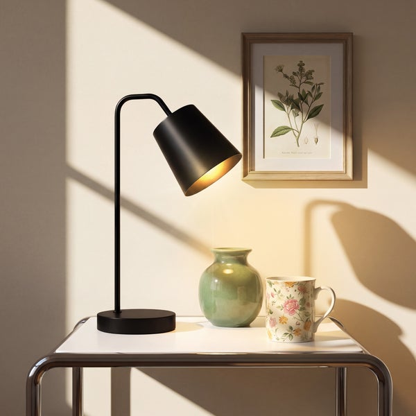 Moderne Schreibtischlampe auf einem Metalltisch neben einer Vase und einem Becher vor einem gerahmten botanischen Bild.