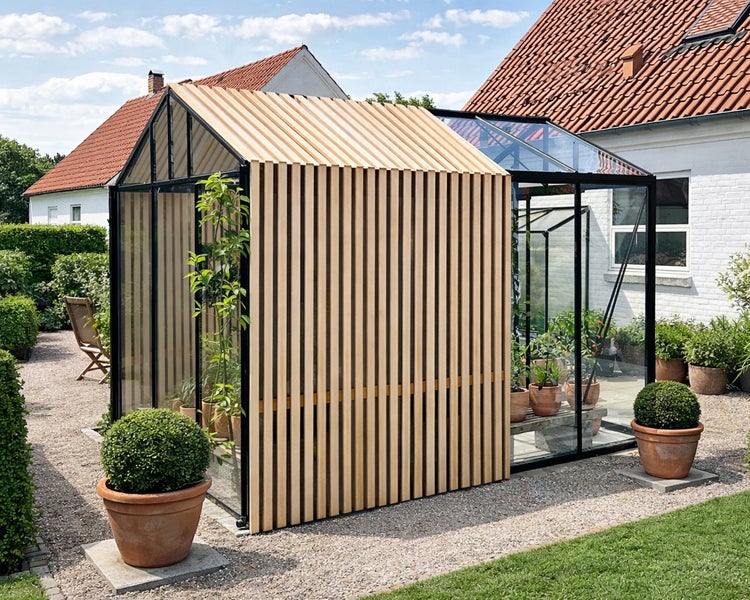 Helles, modernes Gewächshaus mit schwarzem Metallrahmen und Glas, kombiniert mit hellen Holzlamellen, in einer gepflegten Gartenanlage mit Kiesweg.