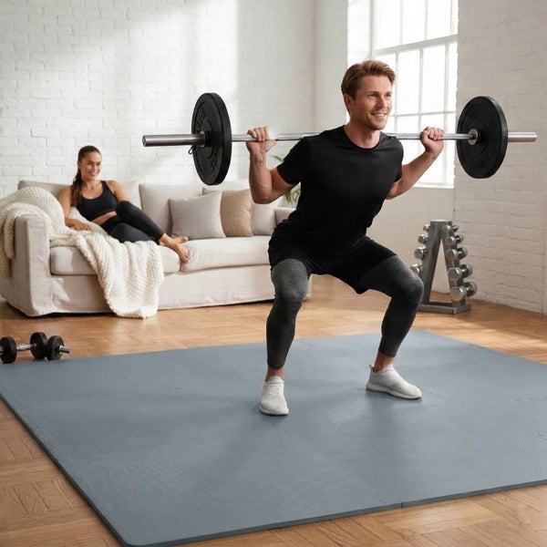 Ein Mann trainiert mit einer Langhantel auf einer grauen Bodenschutzmatte in einem hellen Wohnzimmer mit Sofa und Fitnessgeräten.