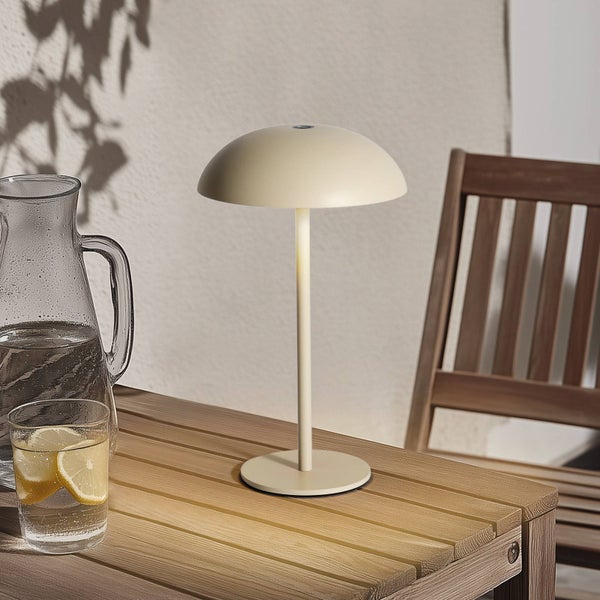 Moderne kabellose LED-Tischleuchte in Beige auf einem Holztisch im Außenbereich, neben einem Wasserkrug und einem Glas mit Zitrone.