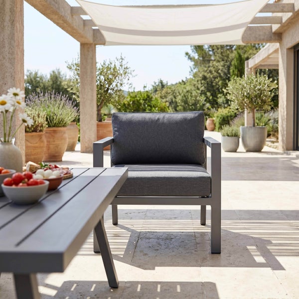 Moderner Garten-Loungesessel mit grauen Polstern und passendem Tisch auf einer hellen Terrasse unter einem Sonnensegel.