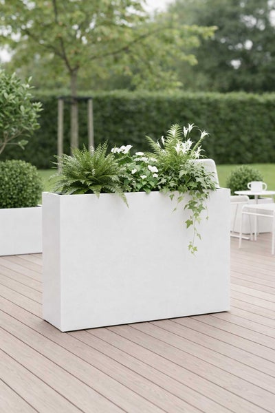 Moderner rechteckiger Pflanzkübel auf einer Holzterrasse, bepflanzt mit Farnen und weißen Blumen vor einer grünen Hecke und Gartenmöbeln.