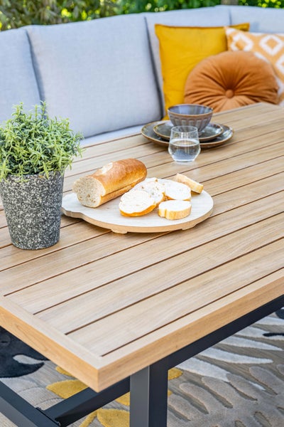 Hell gestalteter Gartentisch mit lamelierter Platte in Holzoptik und Metallgestell vor einem Gartensofa mit Kissen und Dekoration aus Brot und Pflanze