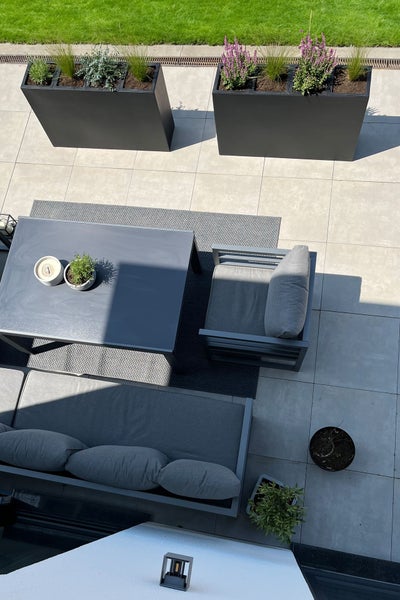 Moderne Terrasse mit grauem Lounge-Set, Tisch auf einem Outdoorteppich und großen rechteckigen Pflanzkästen auf hellen Fliesen.