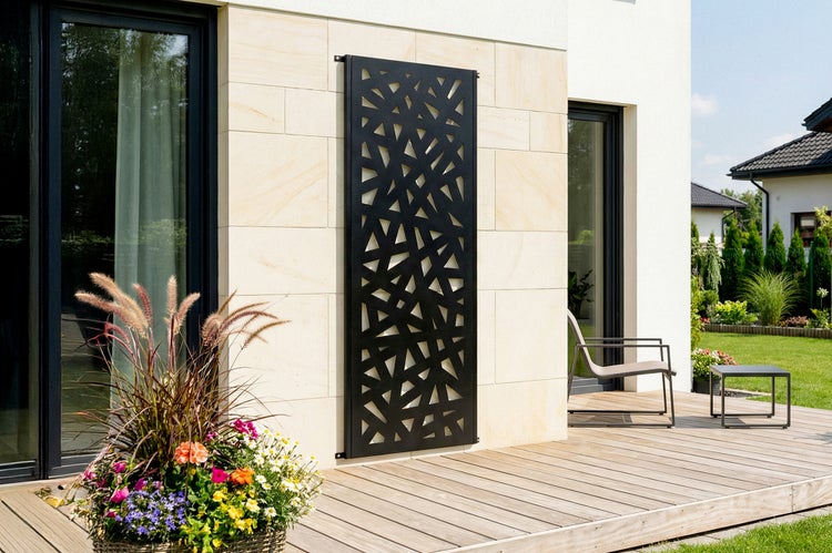Schwarzes dekoratives Wandpaneel aus Metall mit geometrischem Muster an einer hellen Hauswand über einer Holzterrasse mit Blumen und Gartenmöbeln.