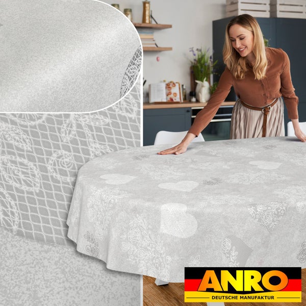 Frau breitet eine ovale Tischdecke mit Herz- und Spitzenmuster in einer hellen Küche aus, ANRO Deutsche Manufaktur Logo.