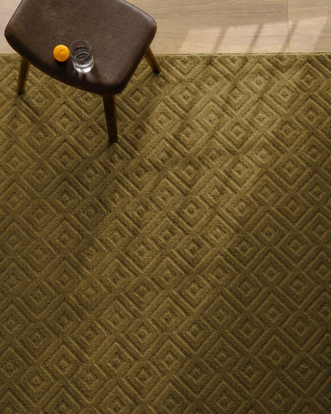 Strukturierter Teppich mit geometrischem Rautenmuster in Reliefoptik, platziert in einem Wohnraum mit einem Hocker auf Holzfußboden.