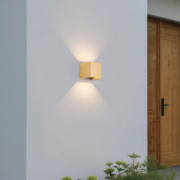 Würfelförmige Außenwandleuchte aus Holz mit Lichtaustritt nach oben und unten an einer Hauswand neben einer Holztür.