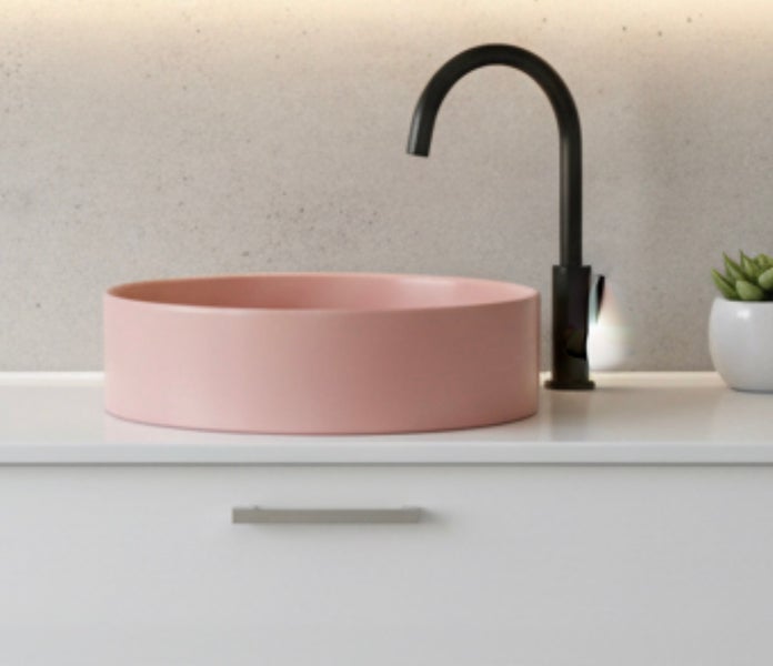 Rosa rundes Aufsatzwaschbecken auf weißem Unterschrank mit schwarzer hoher Armatur vor einer Wand in Betonoptik.